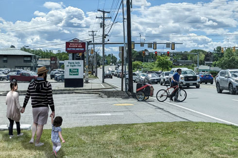 A family holds hands and gets ready to cross a street with high-vehicle traffic at an unsignalized intersection without a crosswalk. A bicyclist with an extended cargo attachment is also trying to cross the busy road perpendicular to traffic.